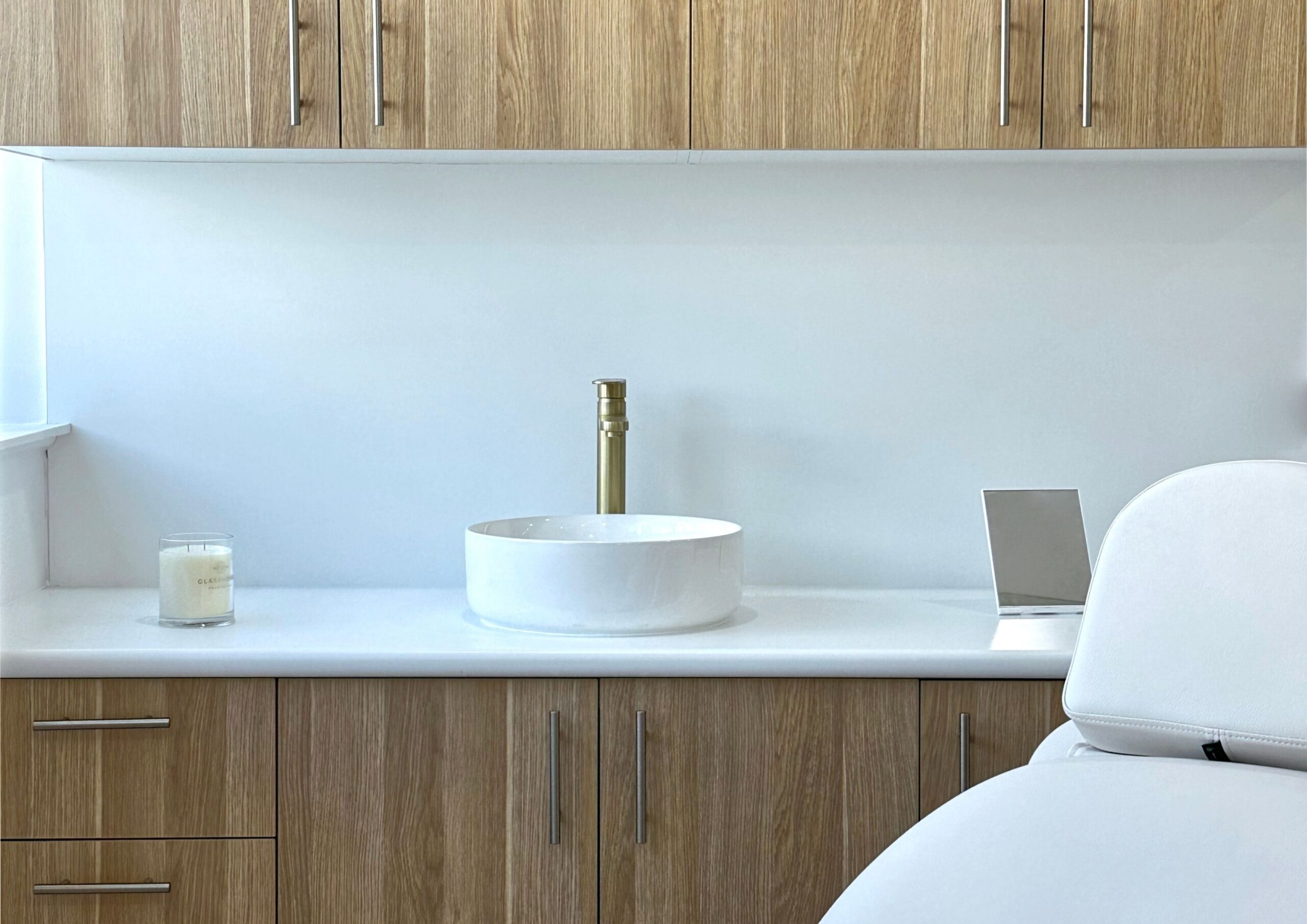 A minimalist bathroom with a round white basin and gold faucet on a white countertop, surrounded by light wood cabinets. A small candle and a square mirror are placed nearby. A white cushioned chair is partially visible in the foreground.