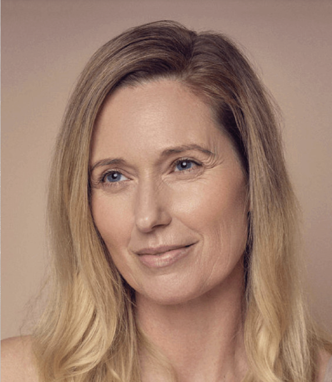 A woman with long, straight blonde hair and blue eyes smiles softly. The background is a neutral beige, emphasizing her natural complexion. She appears calm and relaxed.