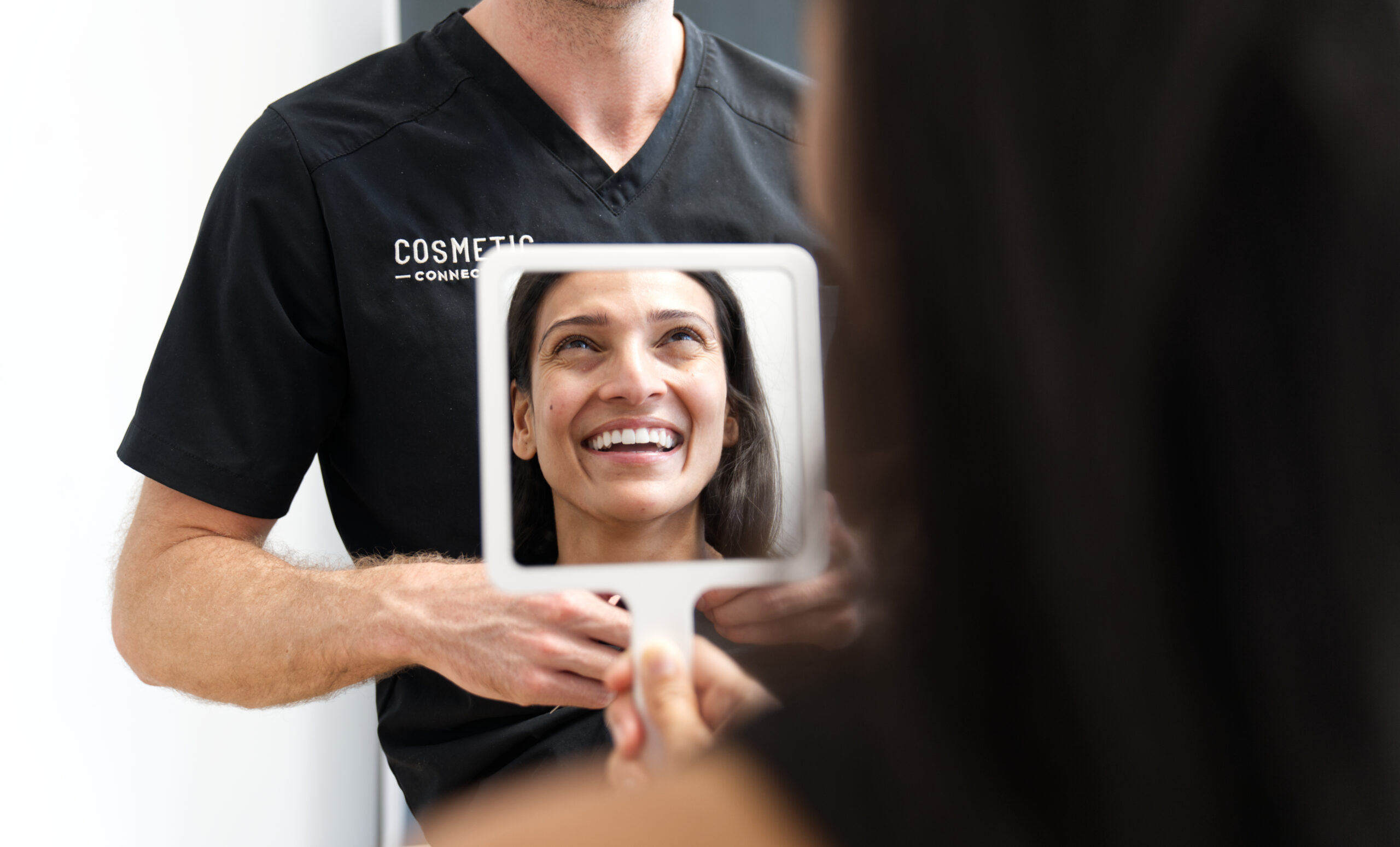 A person wearing a black shirt with Cosmetic printed on it holds a mirror. Another person is seen smiling in the mirrors reflection, creating a joyful expression. The background is softly blurred, focusing on the mirror image.