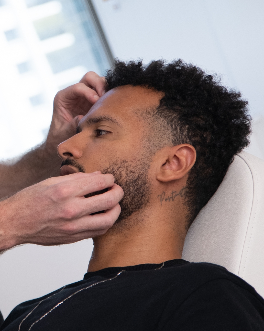 A person with curly hair is seated in a chair while another person examines or adjusts their chin and jawline. The setting appears to be indoors, possibly a clinic or salon.