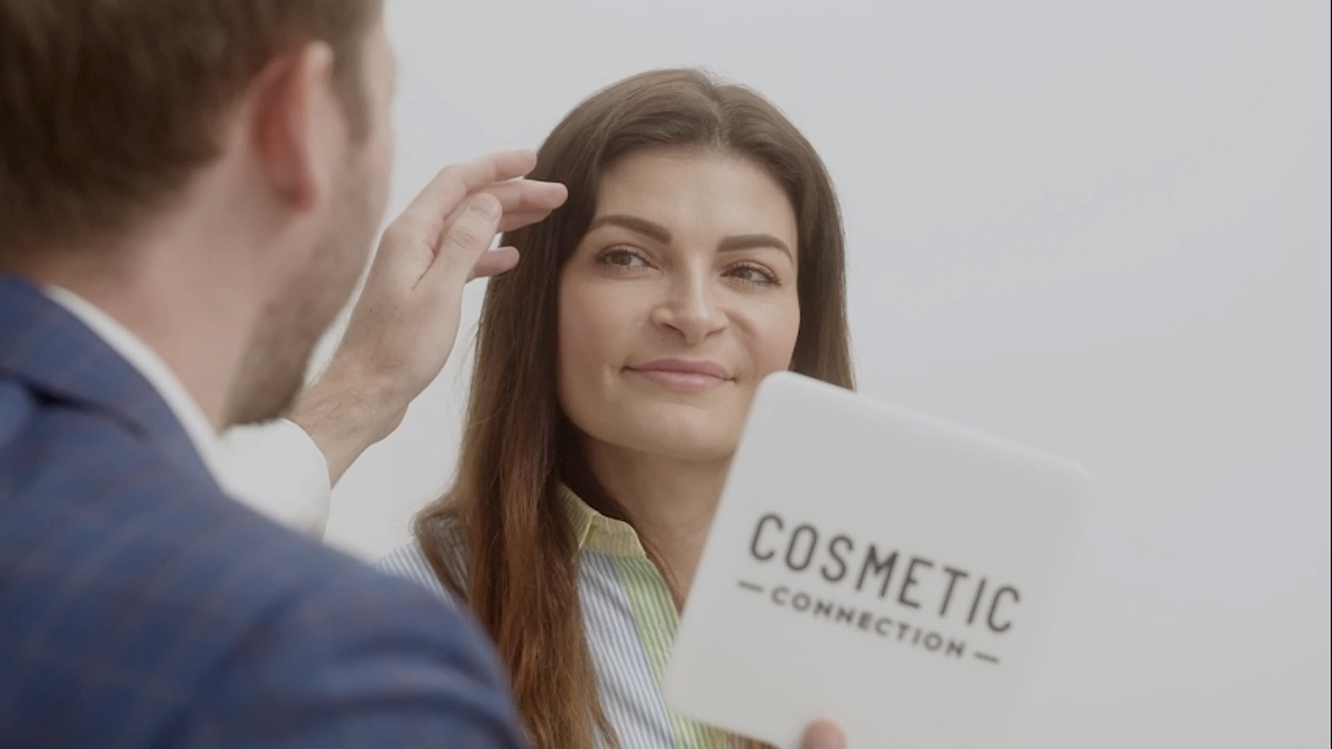 A woman with long hair holds a mirror labeled Cosmetic Connection, as a person gestures near her forehead. She appears to be reviewing cosmetic results in a well-lit setting.