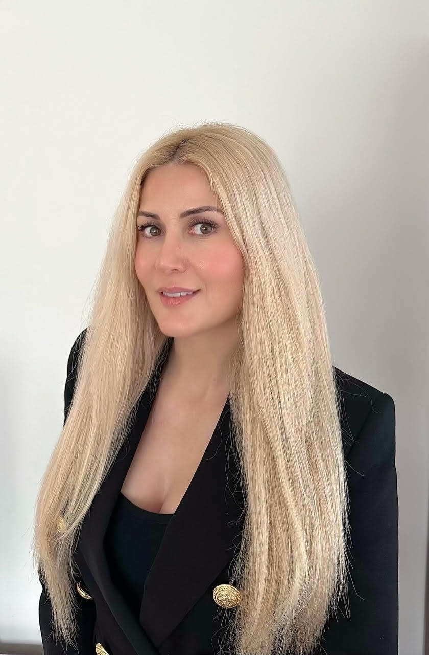 A person with long, straight blonde hair is smiling slightly while facing the camera. They are wearing a black top with gold buttons and standing against a plain white background.