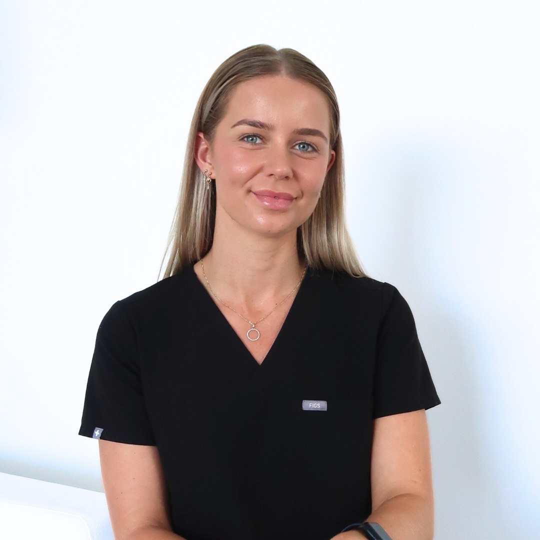 A person with long blonde hair is smiling and wearing a black medical scrubs top. They are standing against a plain white background.