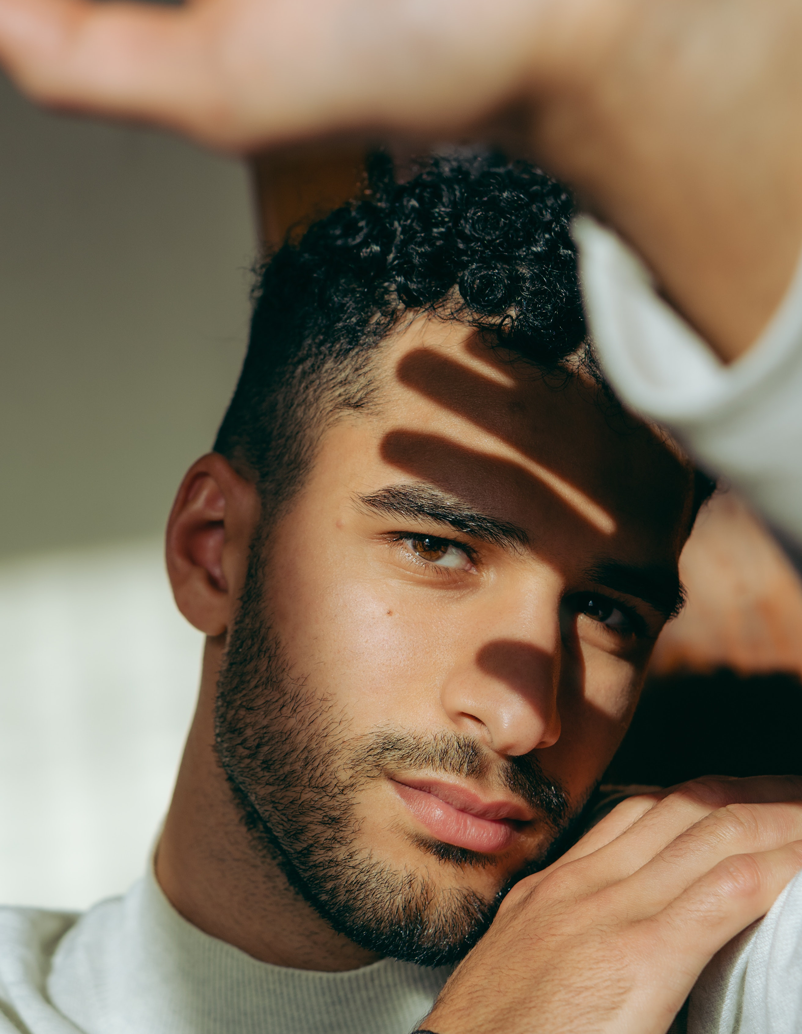 A young man with short curly hair and a trimmed beard gazes into the camera, resting his arm on his shoulder. Sunlight and shadows fall across his face, creating dramatic lines.