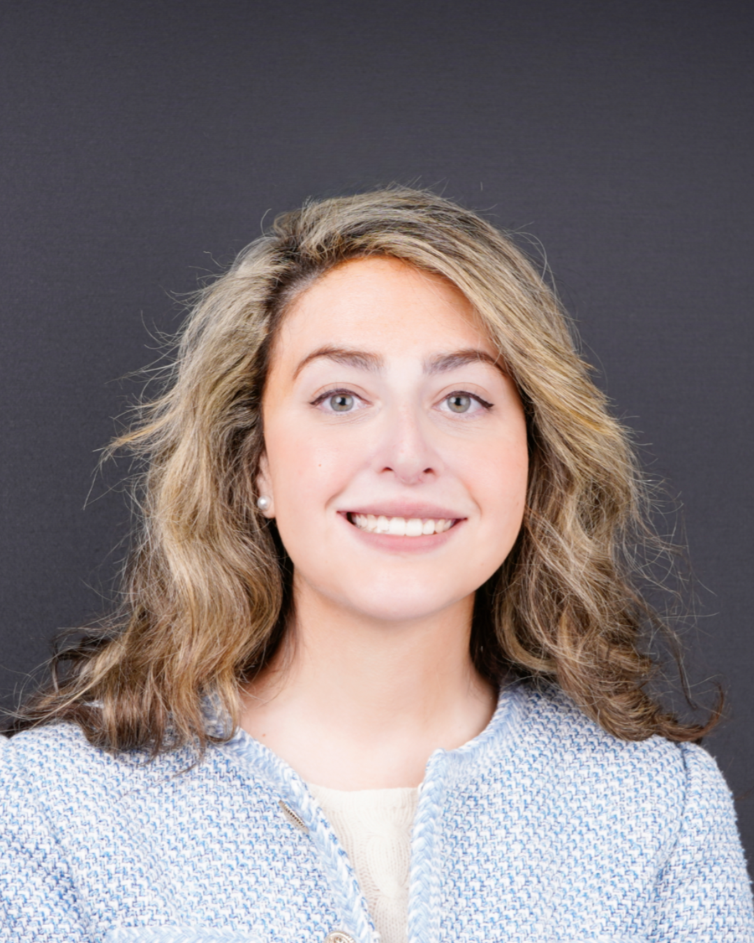 A woman with wavy blonde hair and fair skin smiles at the camera. She is wearing a light blue textured jacket over a cream top, with a dark, plain background behind her.
