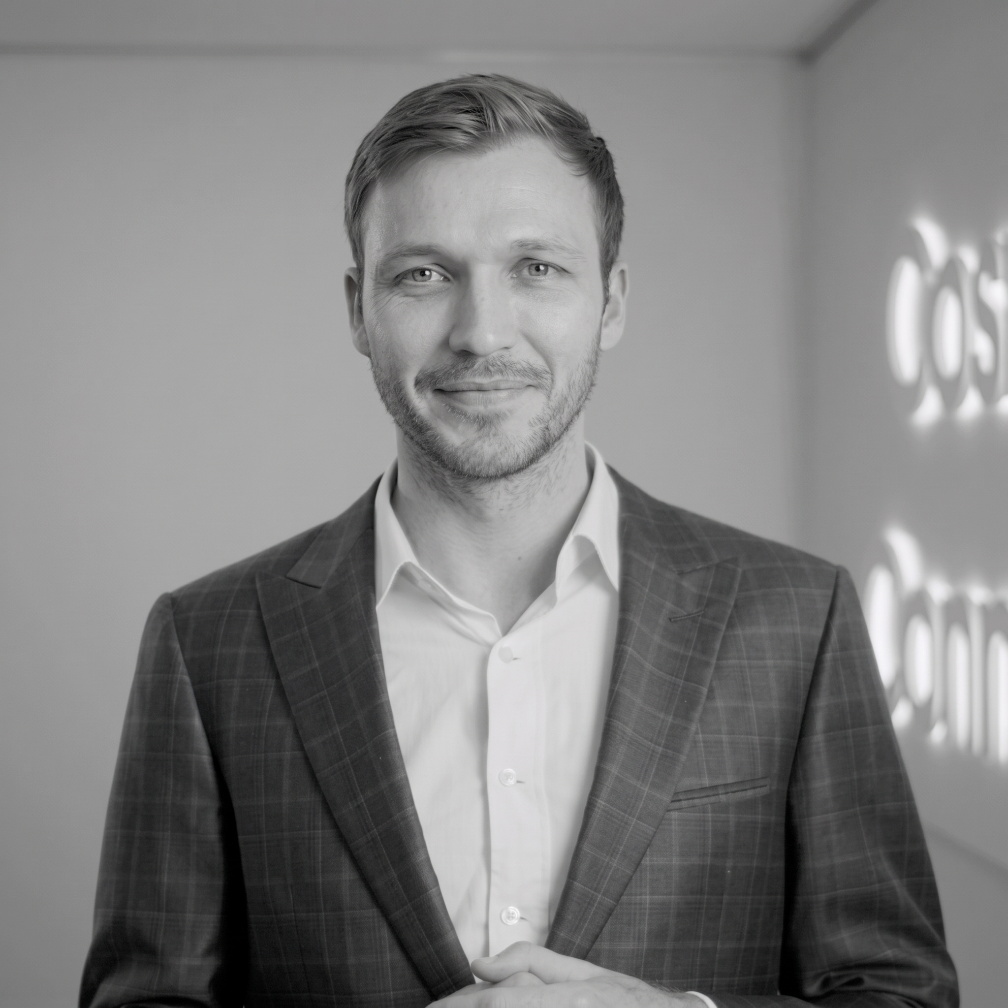 A man in a checked suit jacket and white shirt stands indoors, smiling at the camera. The background is minimal, with blurred white text on the wall.