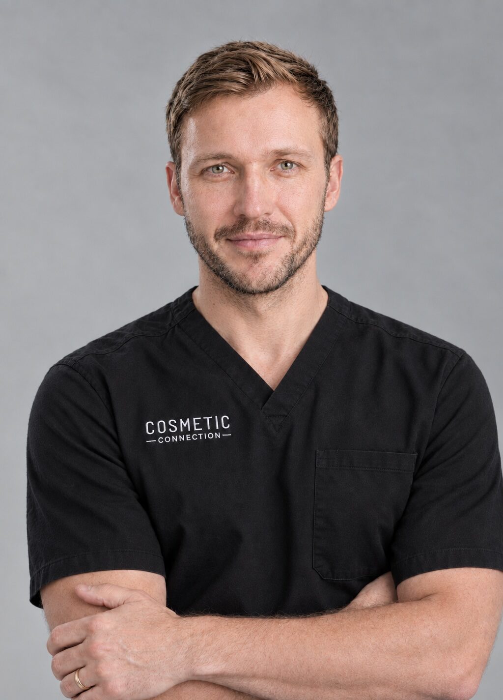 A man with short light brown hair and a beard wears black medical scrubs with Cosmetic Connection on the chest. He stands against a gray background, arms folded, and smiles slightly at the camera.